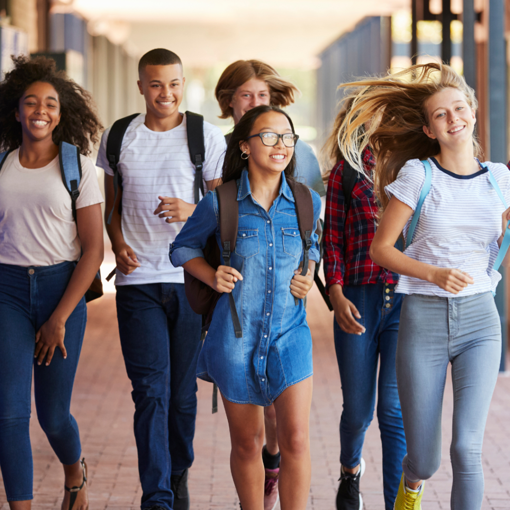 A group of six teenagers walk together down a school corridor, smiling and laughing. They appear energetic and happy as they move forward.