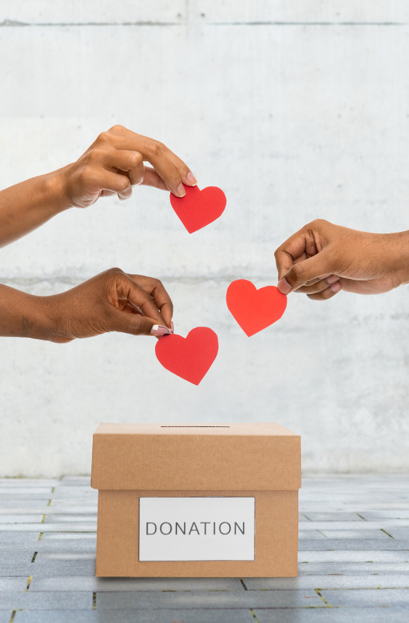 Three hands placing red paper hearts into a cardboard donation box labeled 'DONATION' on a tiled floor, symbolizing giving and generosity."