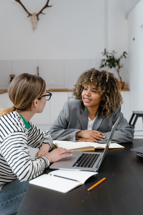 Two women sitting at a table engaged in a discussion, with notebooks and a laptop open in front of them.