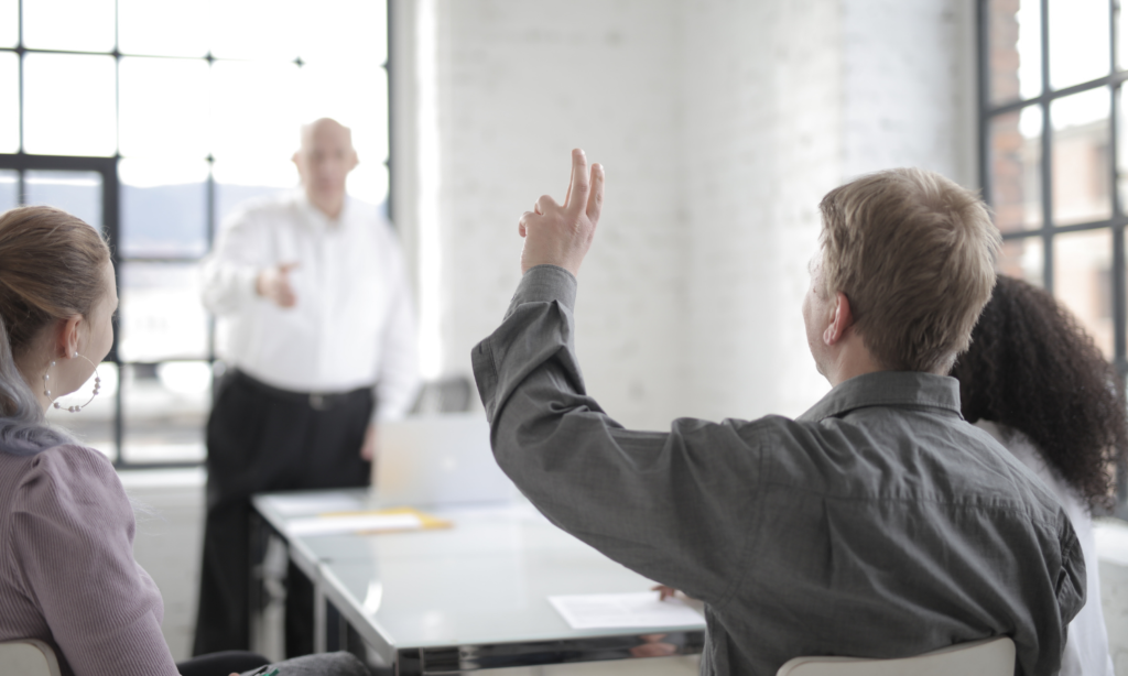 Alt text: Group of people in a bright meeting room listening to a presenter, with one participant raising a hand to ask a question.