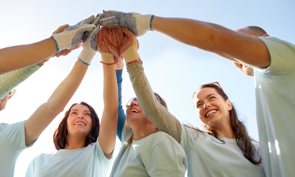 Alt text: Group of smiling volunteers wearing work gloves and light blue shirts, raising their hands together in a team huddle under a bright sky, symbolizing unity, teamwork, and community service.