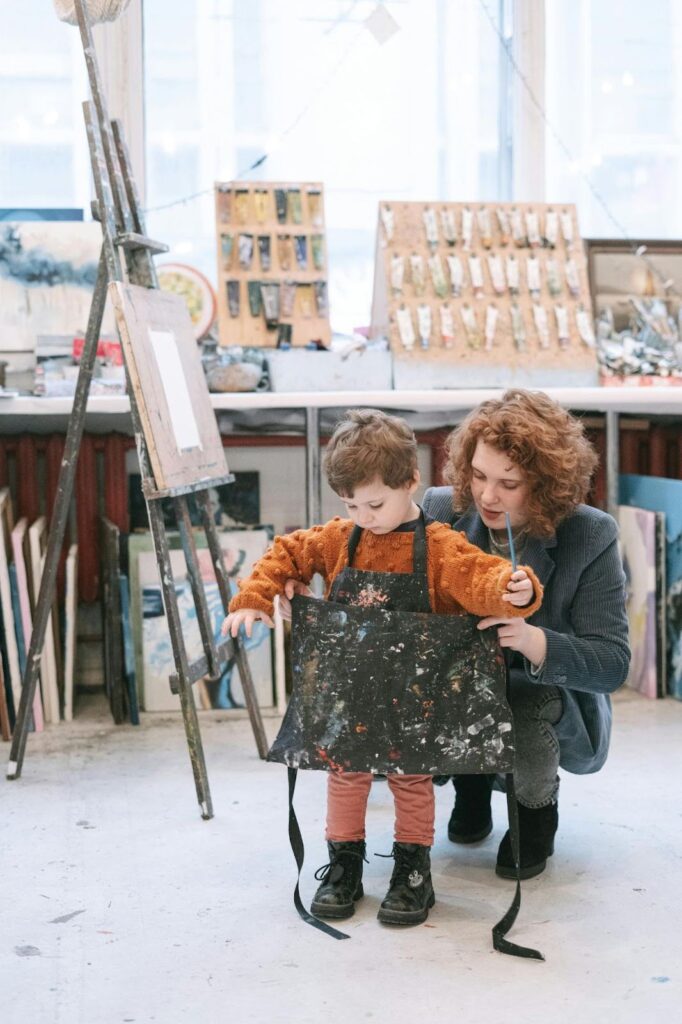 Alt text: Woman helping a young child put on a painting apron with art supplies and an easel in the background.