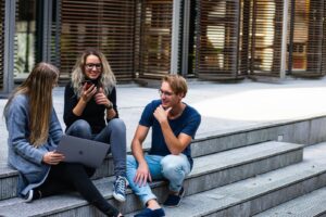 Three young adults sitting on a building’s steps, holding a laptop, a phone, and talking to each other.