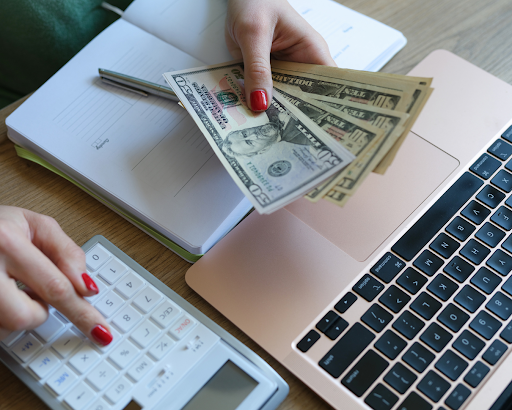 someone typing into a calculator with money in their hands. A notebook and laptop are sitting on the table underneath