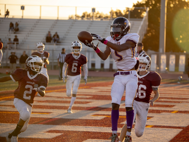 High school football player in white jersey catching a pass during a game at sunset, with other players and stadium bleachers visible in the background