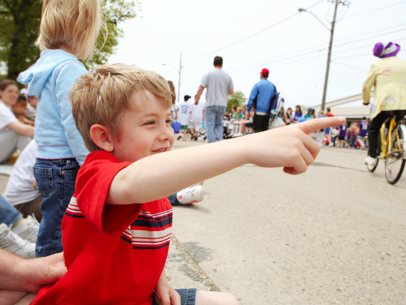 Young boy in a red shirt sitting on the curb and pointing excitedly as a parade passes by, with other children and families watching nearby.