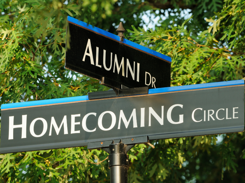 Street sign intersection showing 'Alumni Dr' and 'Homecoming Circle' with green foliage in the background
