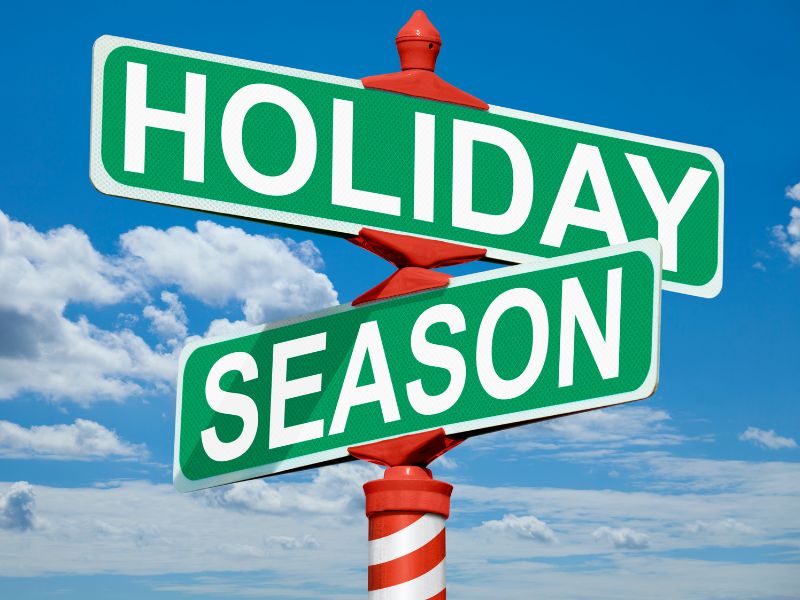 Green street signs reading 'Holiday' and 'Season' intersecting on a red and white striped pole against a blue sky with white clouds