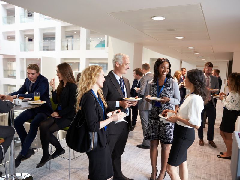 Professional networking event in modern office atrium with business professionals in formal attire socializing with plates of food