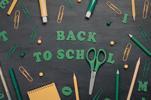 Flat lay of green and gold school supplies including pencils, paperclips, scissors, and notepad arranged around 'Back to School' letters on chalkboard backgroun