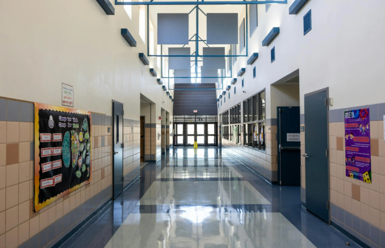 Empty school hallway with high ceilings, bulletin boards, and sunlight reflecting on polished floors.