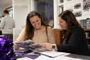 Two adult women smiling and reviewing printed materials together at a table during a school community event, creating a warm and collaborative atmosphere in a public school setting.