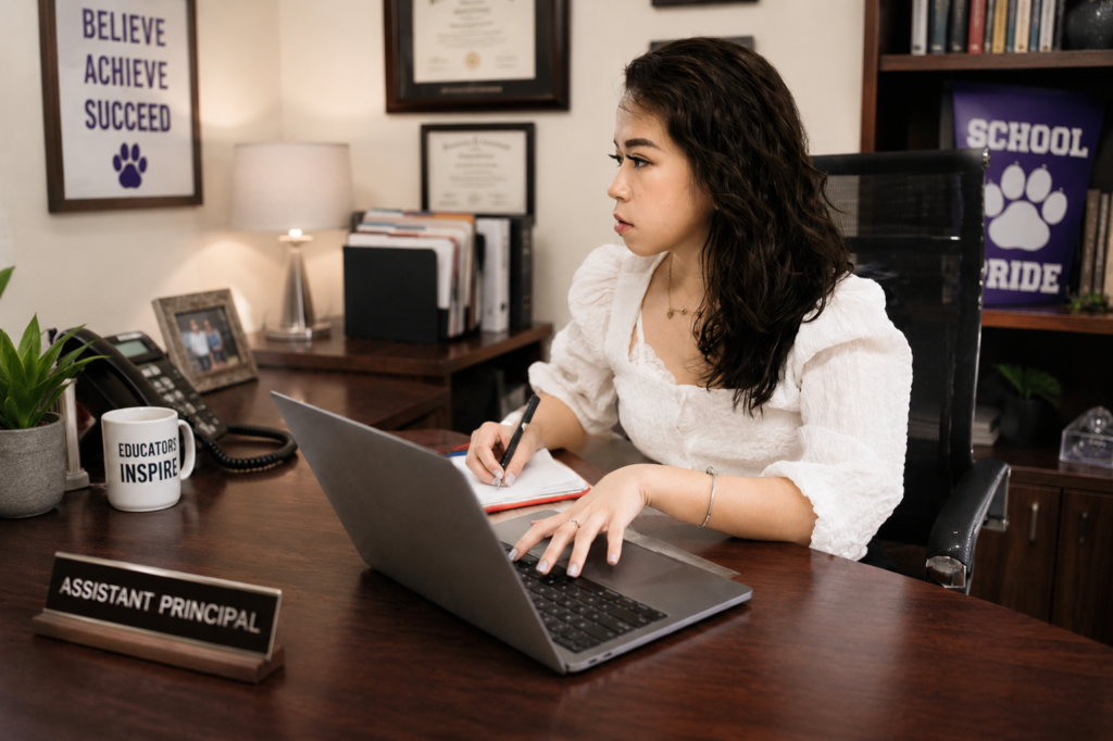 School administrator working at a desk in a professional school office, using a laptop and taking notes with motivational school decor, framed certificates, and educational materials visible in the background.
