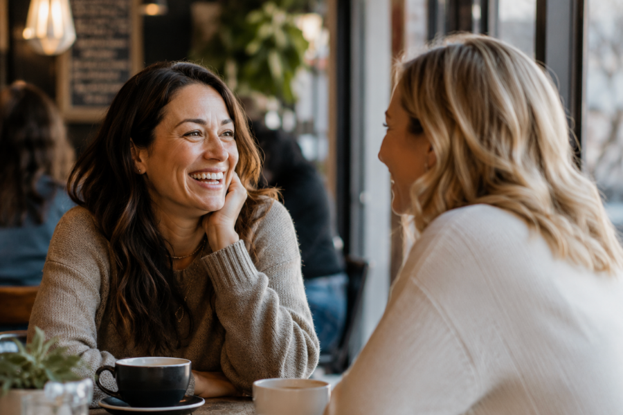 Two adult women laugh and talk across a coffee shop table, sharing a warm and genuine conversation in a cozy community setting.